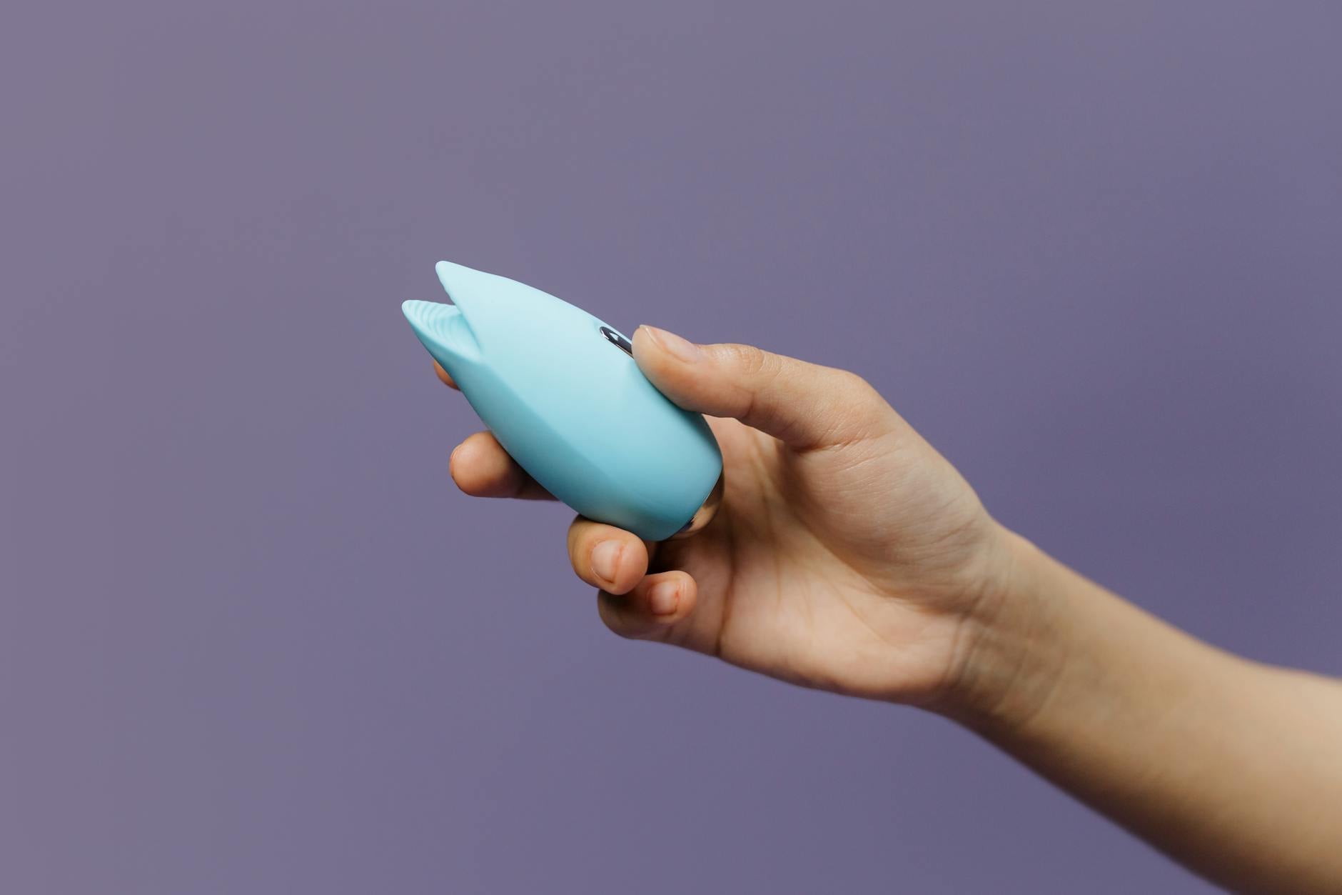 A hand holding a sleek blue clitoral vibrator against a solid purple background